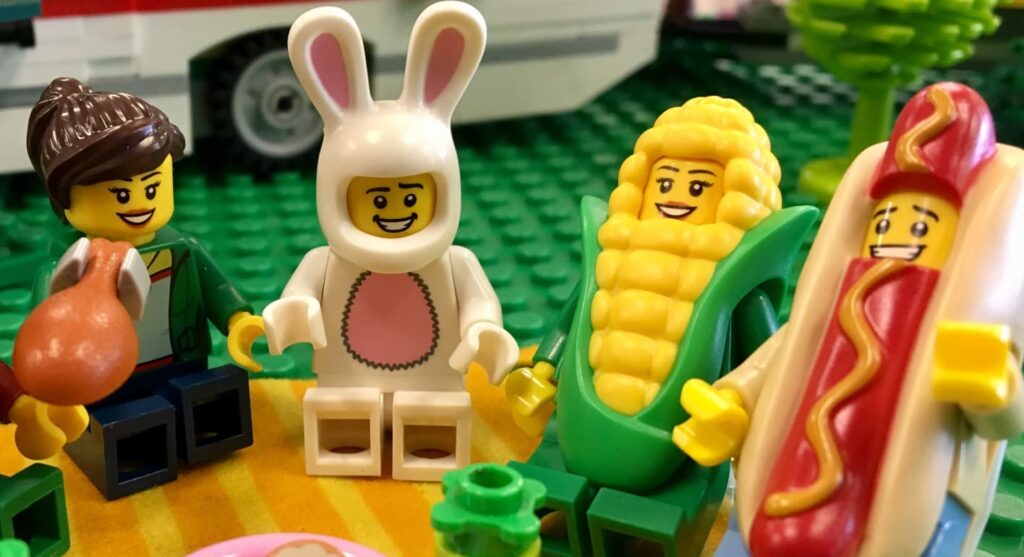 LEGO mini-figures sitting together, dressed in fun costumes including a bunny suit, corn cob outfit, and hot dog suit, enjoying a picnic.