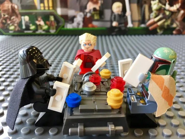 LEGO mini-figures, including Darth Vader, Boba Fett, and others gathered around a table with colorful pieces, appearing to play a board game