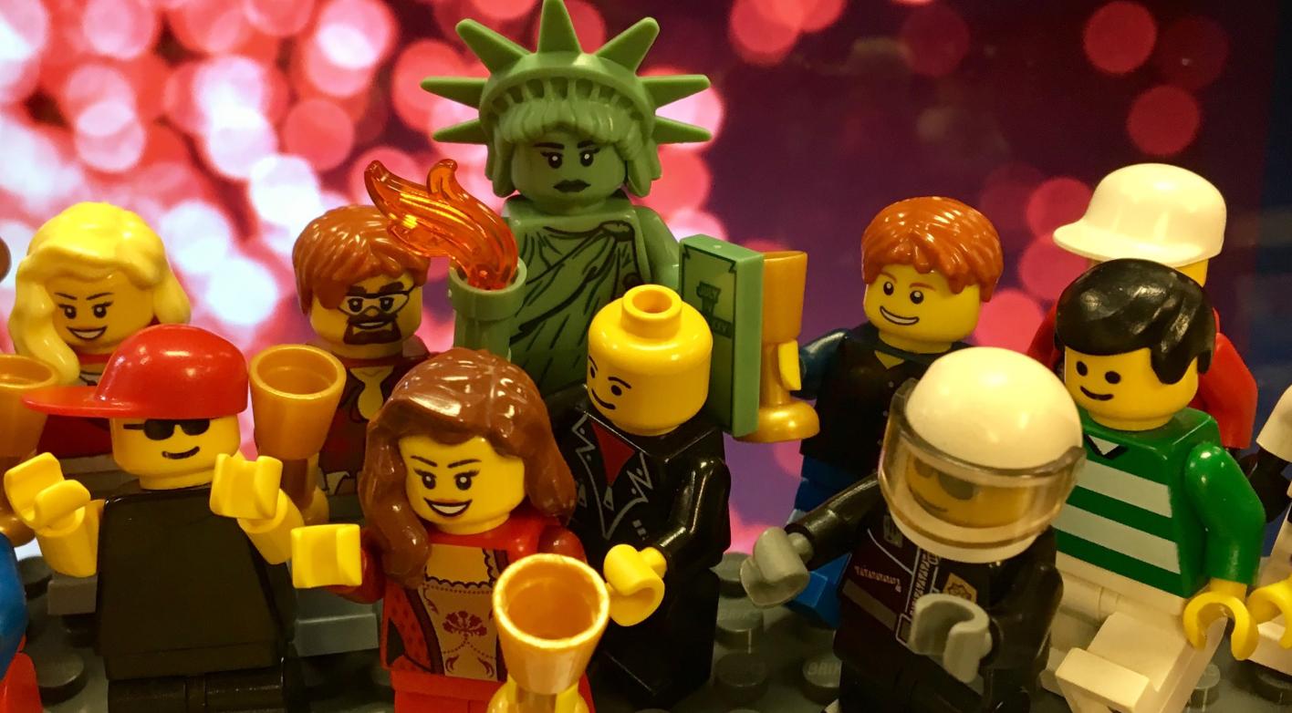Group of LEGO mini-figures celebrating, including a Statue of Liberty figure, various characters holding cups, and colorful background lights