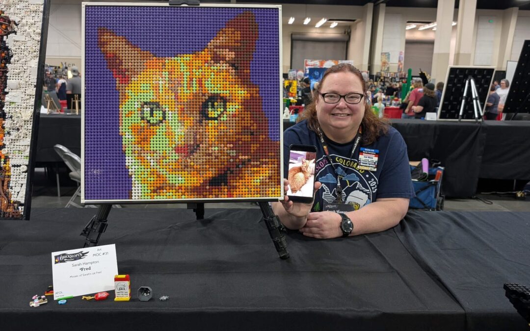 Sarah sits at a LEGO show holding an image of her cat on her phone with a LEGO mosaic of him she built is next to her.