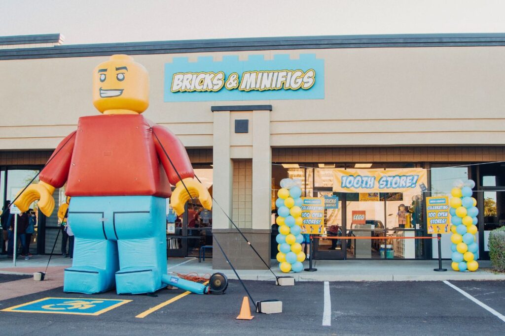 Large inflatable LEGO minifig outside a Bricks & Minifigs store that has a banner announcing that it is the 100th store to open and has yellow and blue balloons outside. 