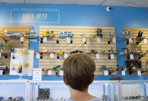A boy looks at a wall of LEGO® sets behind a counter. A sign reading "Used Sets" is on the wall above it.