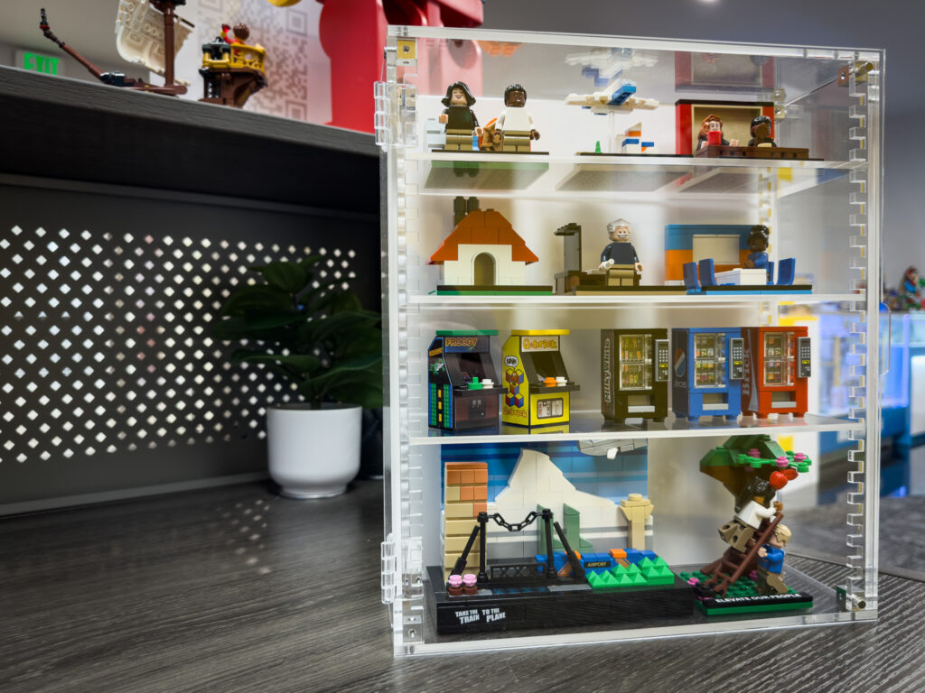 An clear display case holding tiny LEGO builds, including arcade machines, vending machines, scenery, and a few minifigs.