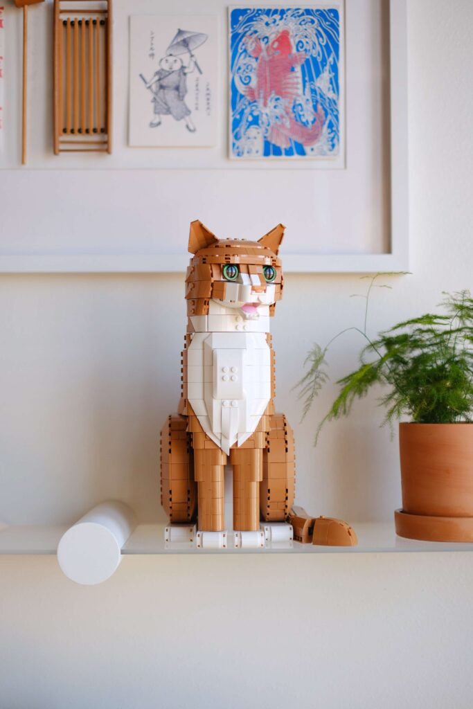 The Orange Cat LEGO® set on a shelf beneath paintings and next to a potted plant.